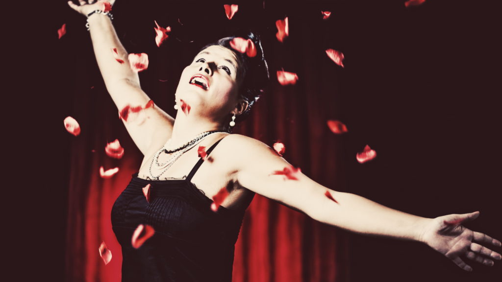 A woman performing on stage, arms outstretched with rose petals falling around her, set against a dark red background. This image symbolises artistic expression and performance. Having a clear out or selling online? From craftwork to Kraftwerk, HMRC may soon know—a reminder for performers that selling goods or crafts could have tax implications.